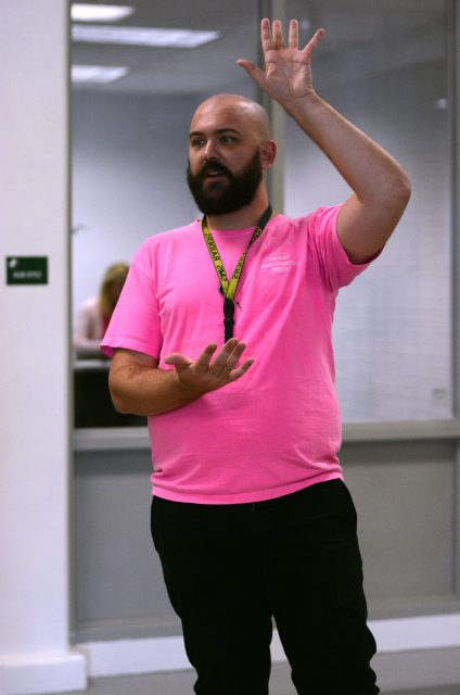 Rucker stands at the front of a classroom with one hand up. He appears to be giving instructions.