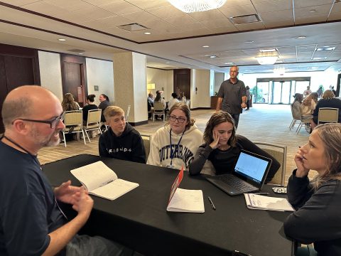 An image of a teacher and studenst seated together at a table covered with various papers and documents, engaged in a collaborative learning session.